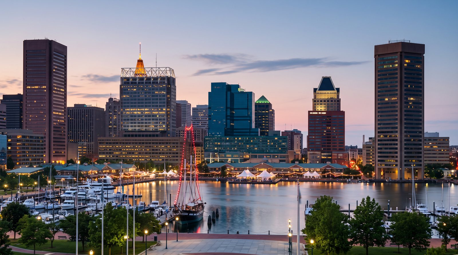 The Baltimore skyline across the Inner Harbor at twilight, with a tall ship lit in red, waterfront pavilions and the towers reflected in the basin